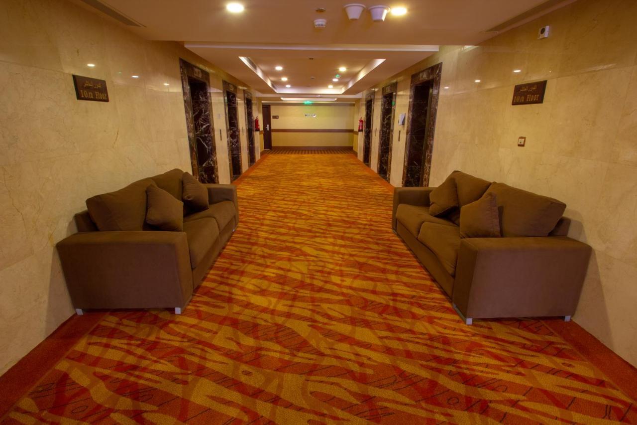 Grand Makkah Hotel La Mecque Extérieur photo The photo shows a hallway inside a building, likely a hotel. There are two brown sofas placed opposite each other on either side of the corridor. The floor features a patterned carpet with warm colors, and the walls are finished with a smooth, light-